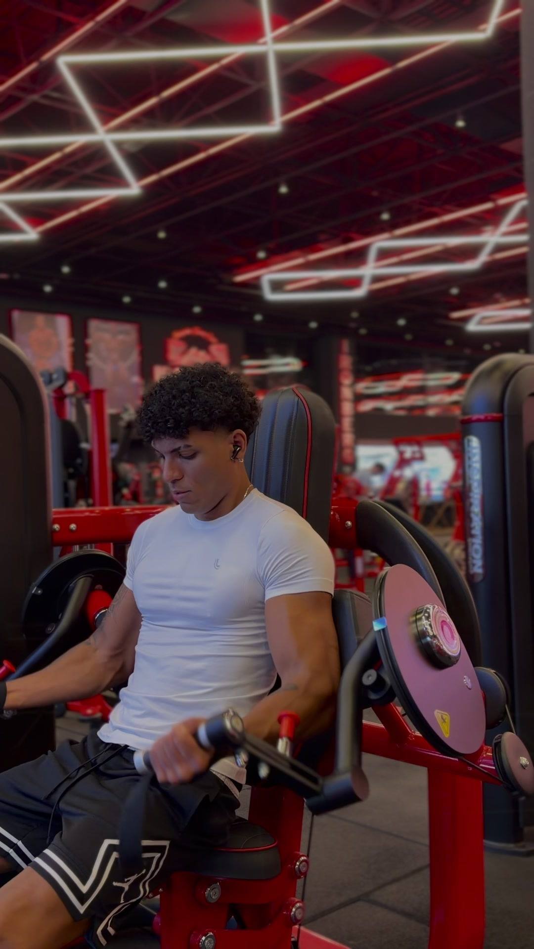 Matheus Souza treinando bíceps na máquina, em uma academia com luzes de neon